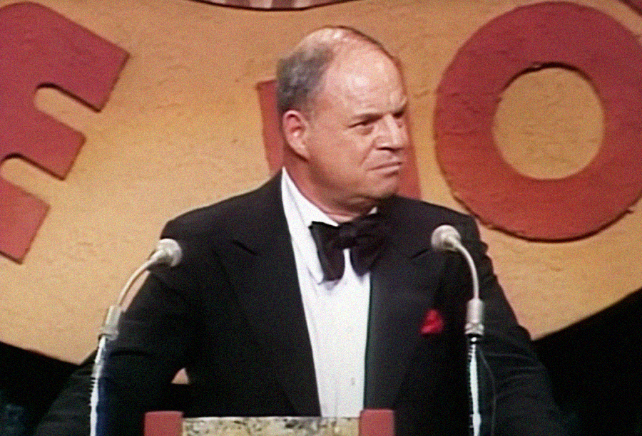 A man in a tuxedo and bow tie stands at a podium with two microphones, in front of a large sign with bold red letters on a tan background. He has a serious expression on his face.