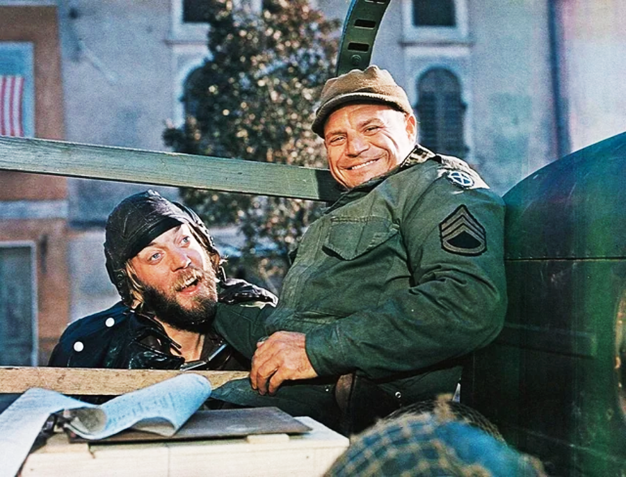 Two men in military gear sit smiling in an open vehicle. One wears a helmet and goggles, making a funny face, while the other, in a green jacket with sergeant stripes, grins. An American flag hangs in the blurred background.