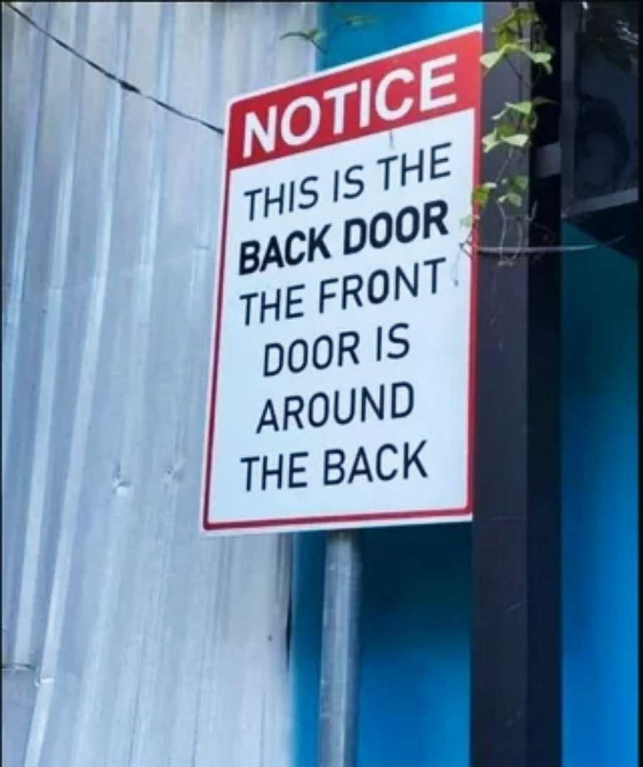A sign reads: "NOTICE. THIS IS THE BACK DOOR. THE FRONT DOOR IS AROUND THE BACK." The sign is posted on a wall next to a metal siding and some plants.
