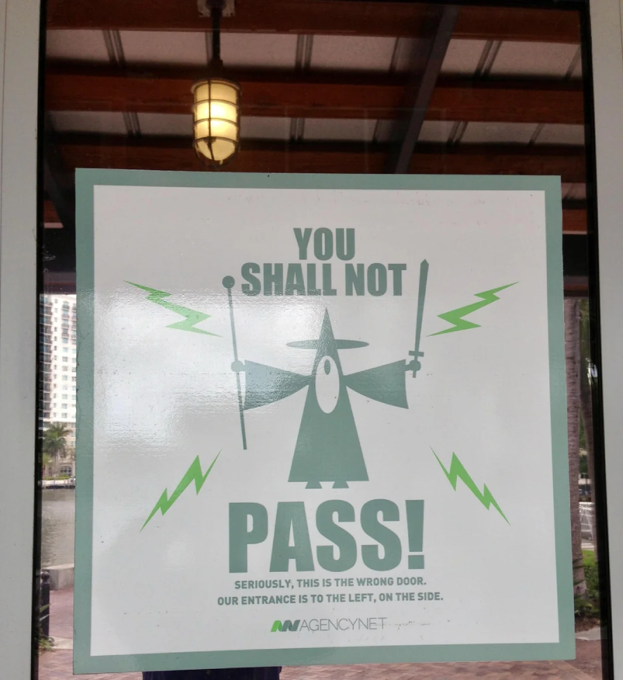 A sign on a glass door shows a wizard holding a staff and rod, with green lightning bolts and the text: "You shall not pass! Seriously, this is the wrong door. Our entrance is to the left, on the side.