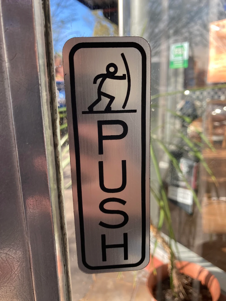 A metal sign on a glass door shows a stick figure pushing against a vertical line, with the word "PUSH" written vertically below the figure.