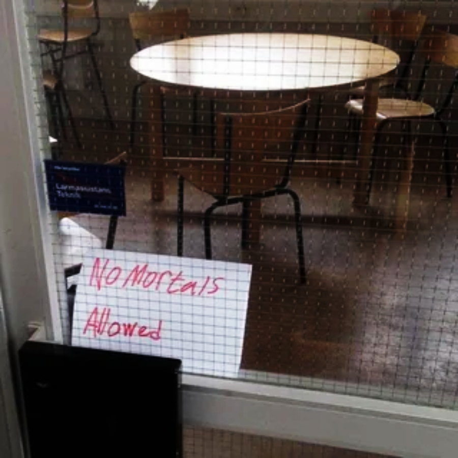 A round table with wooden chairs in an empty room, viewed through a glass door. A handwritten sign on the door reads, "No Mortals Allowed" in red marker.