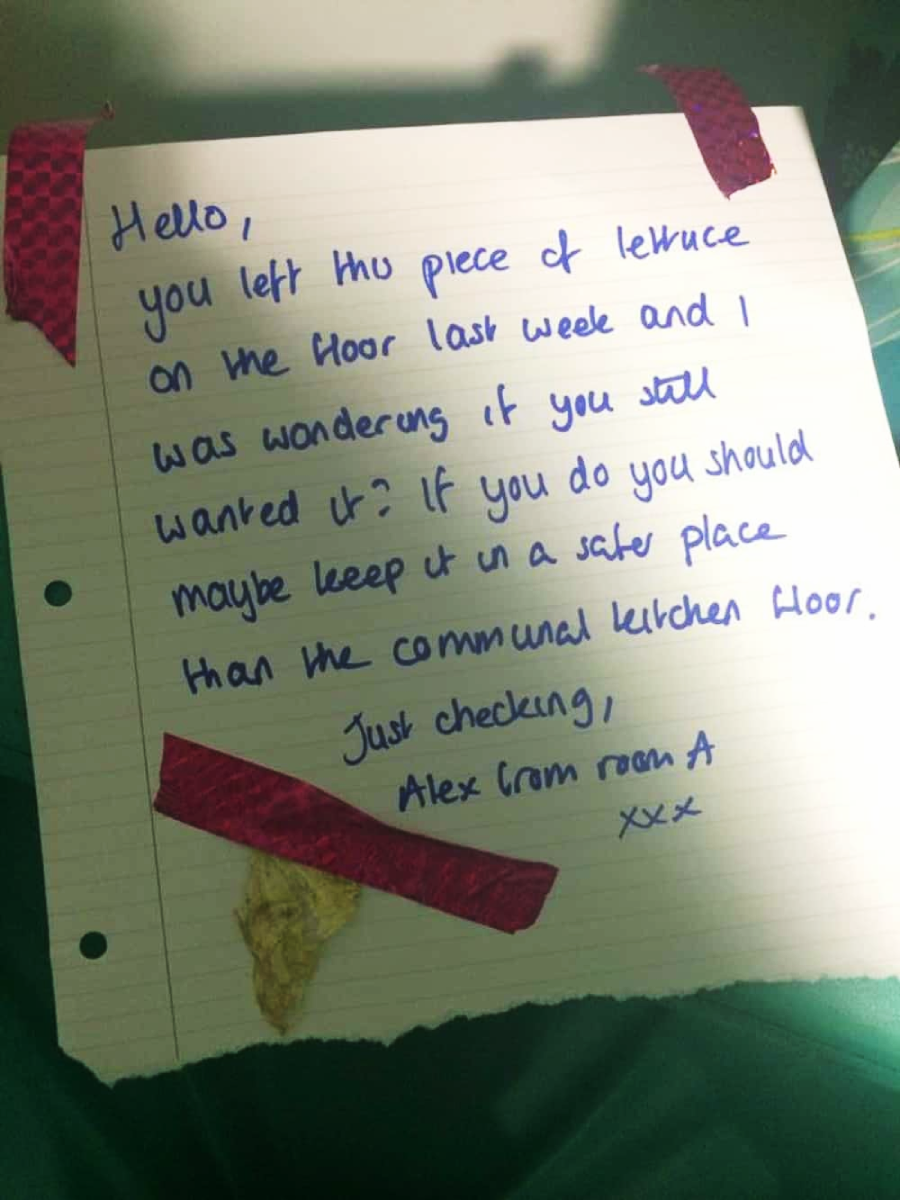 A handwritten note on lined paper, taped with red patterned tape, reads: "Hello, you left this piece of lettuce on the floor last week and I was wondering if you still wanted it? If you do, you should maybe keep it in a safe place than the communal kitchen floor. Just checking, Alex from room A xxx." A dried lettuce piece is taped to the note.
