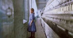 A young girl in a blue dress stands alone beside large textile machinery in a mill, gazing out a window. The scene is lit by daylight streaming in from the windows, highlighting the industrial setting.