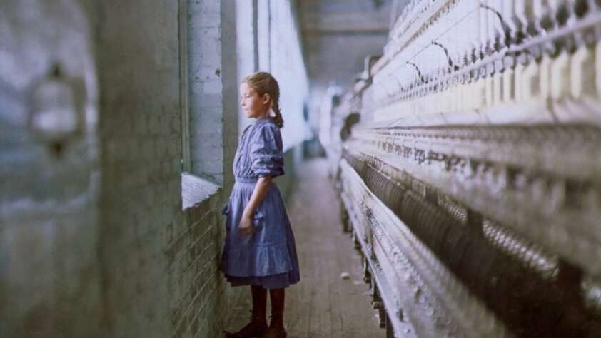 A young girl in a blue dress stands alone beside large textile machinery in a mill, gazing out a window. The scene is lit by daylight streaming in from the windows, highlighting the industrial setting.