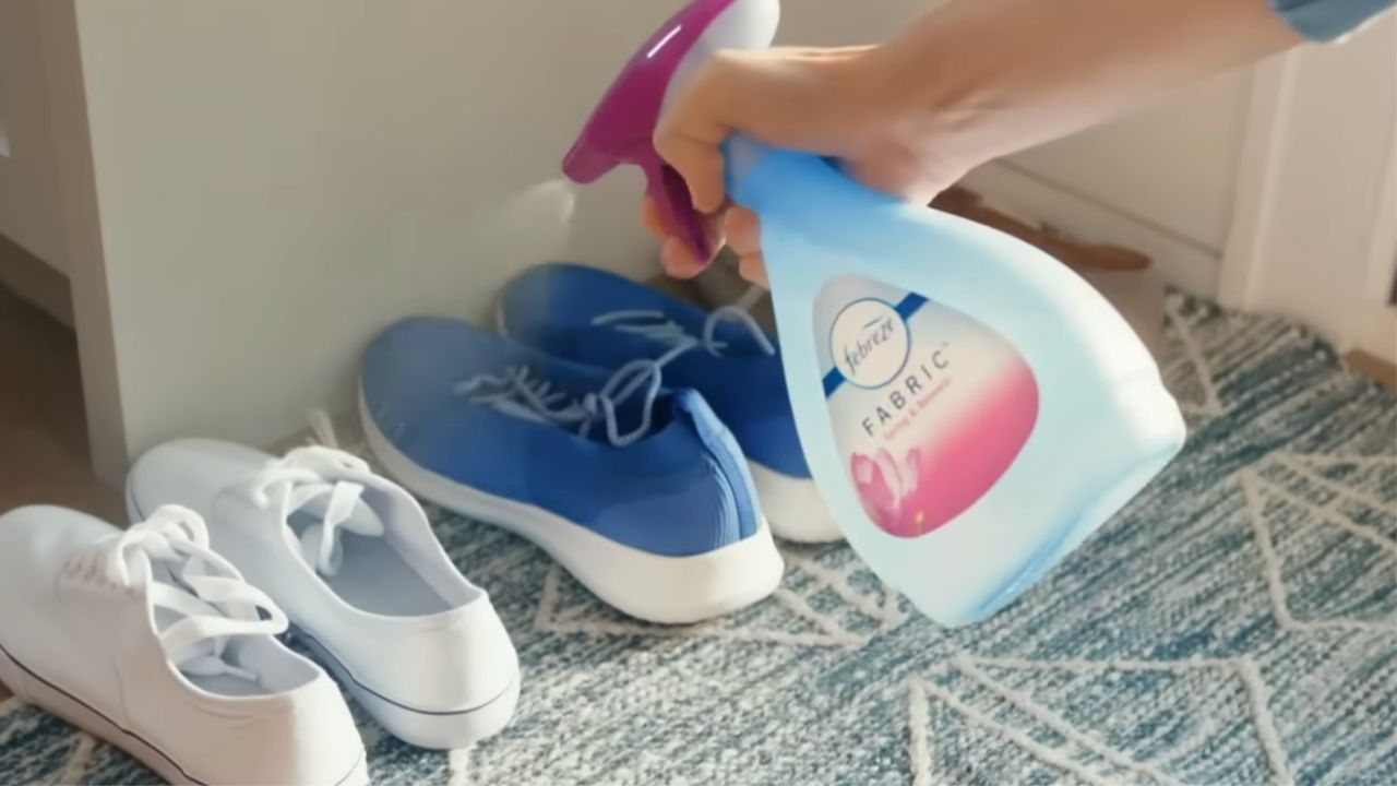 A hand sprays Febreze Fabric Refresher onto blue sneakers next to white shoes on a patterned rug by a cabinet.