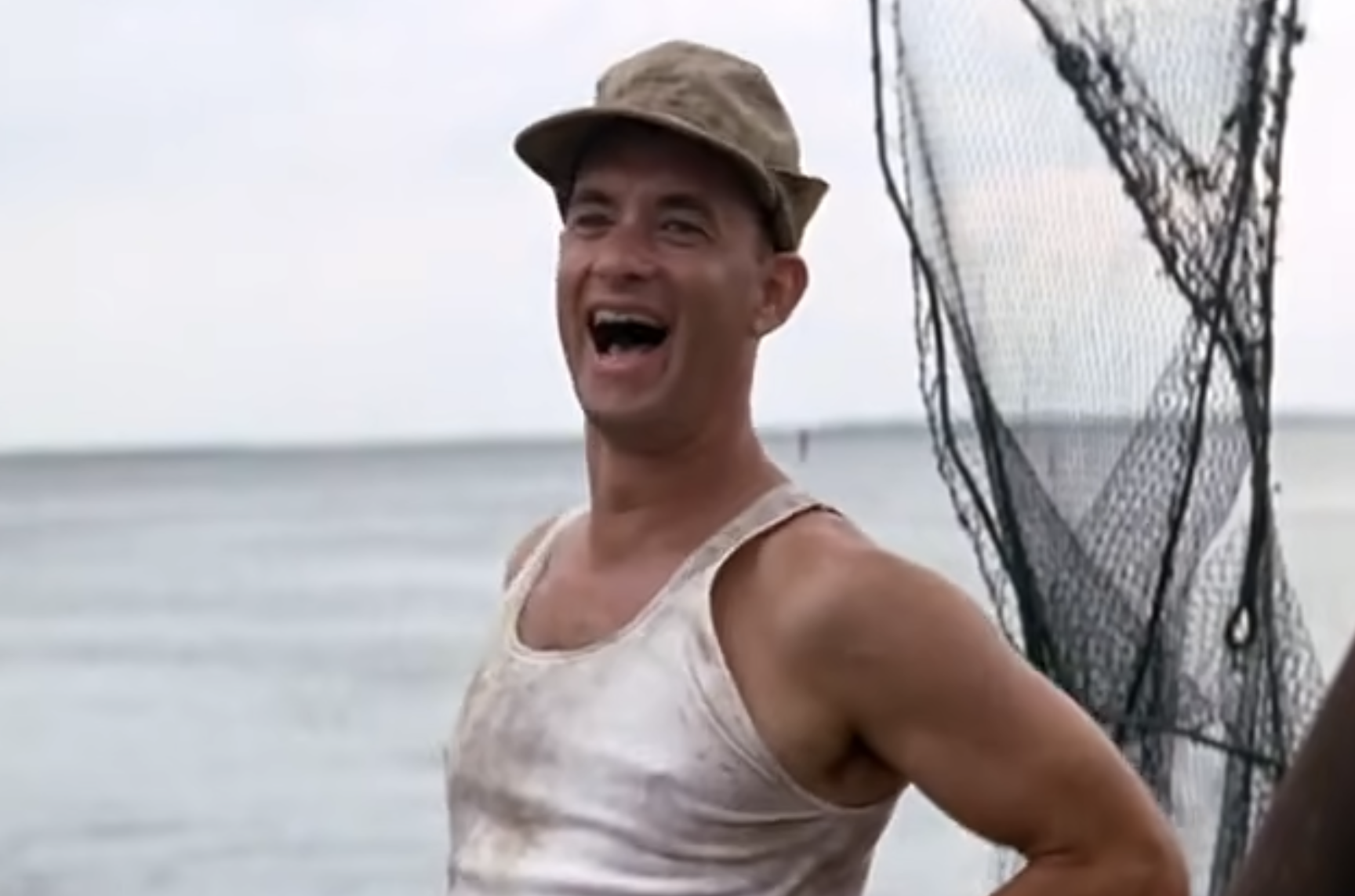 A man in a dirty tank top and cap smiles broadly while standing near fishing nets by the water.