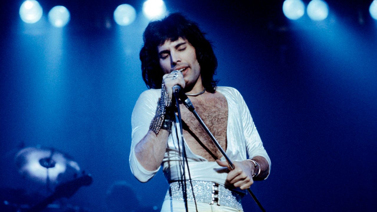 A singer in a white, deep V-neck outfit performs on stage under blue lights, passionately holding a microphone.