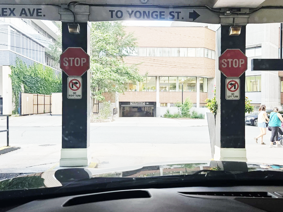 View from a car exiting a parking garage, showing two stop signs, no left turn and no right turn signs, and a street with a few pedestrians in the background.
