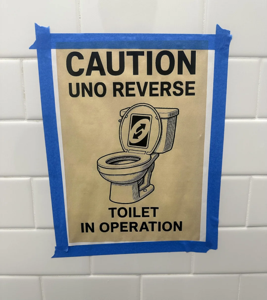 A sign taped to a tiled wall reads "CAUTION UNO REVERSE TOILET IN OPERATION" with an illustration of a toilet and a reverse arrow symbol on the lid, referencing the UNO reverse card.