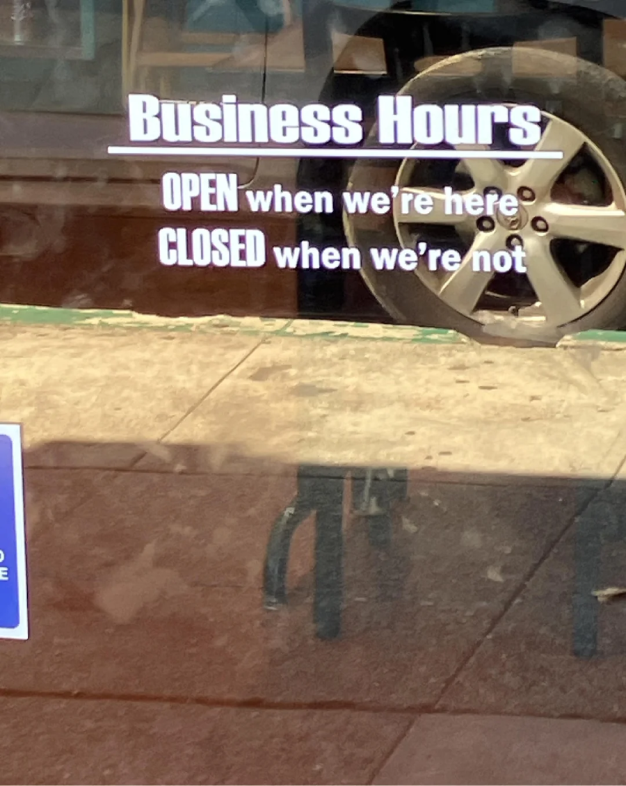 A glass door displays a humorous sign reading: "Business Hours: OPEN when we're here, CLOSED when we're not." A car wheel and sidewalk are reflected in the glass.