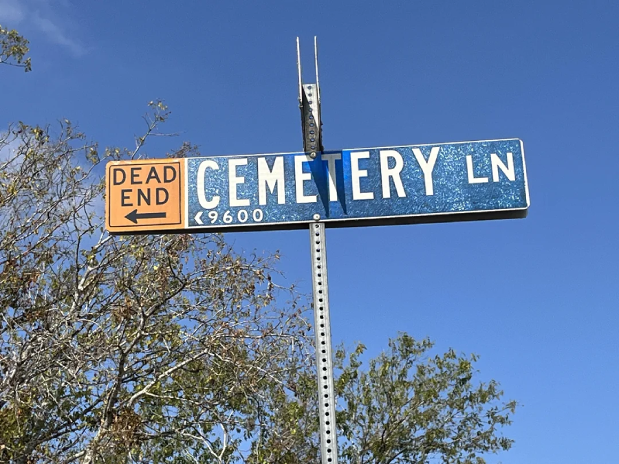 A street sign reads "Cemetery Ln 9600" with a smaller sign above it that says "Dead End" and points left, set against a blue sky and some leafy trees.