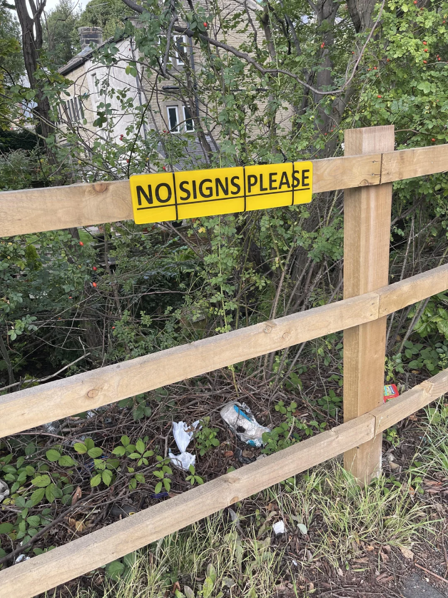 A yellow sign on a wooden fence reads "NO SIGNS PLEASE." Bushes, litter, and a house are visible in the background.