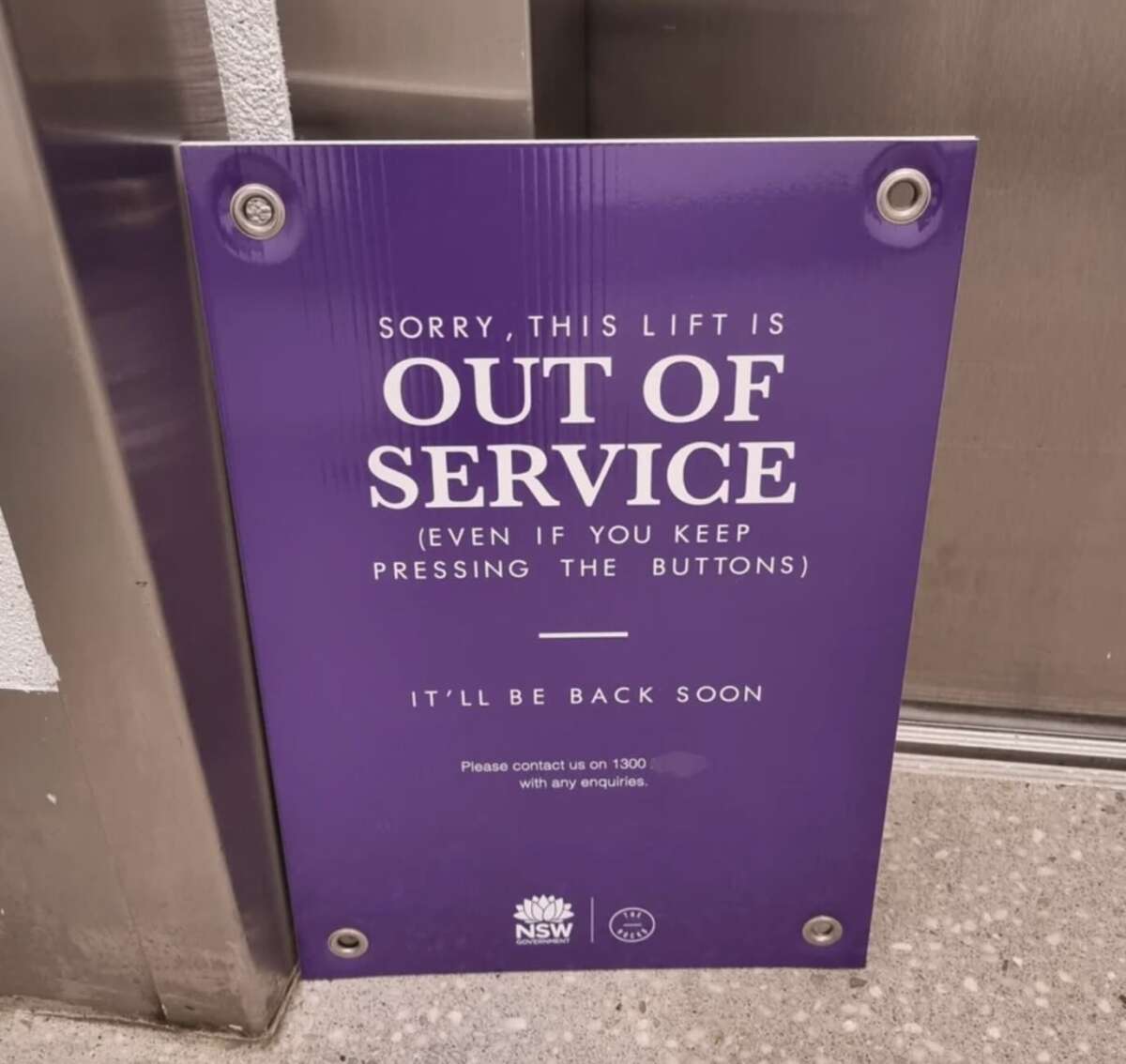 A purple sign leaning against a wall reads: "Sorry, this lift is OUT OF SERVICE (even if you keep pressing the buttons). It'll be back soon. Please contact us on 1300 with any enquiries.