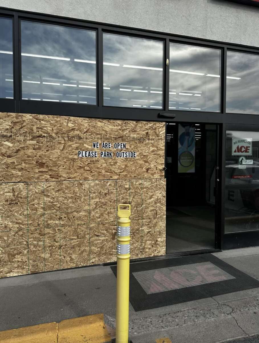 A storefront with boarded-up windows displays a sign that reads, “WE ARE OPEN PLEASE PARK OUTSIDE.” The entrance door is glass, and a yellow pole stands near the entrance on the sidewalk.