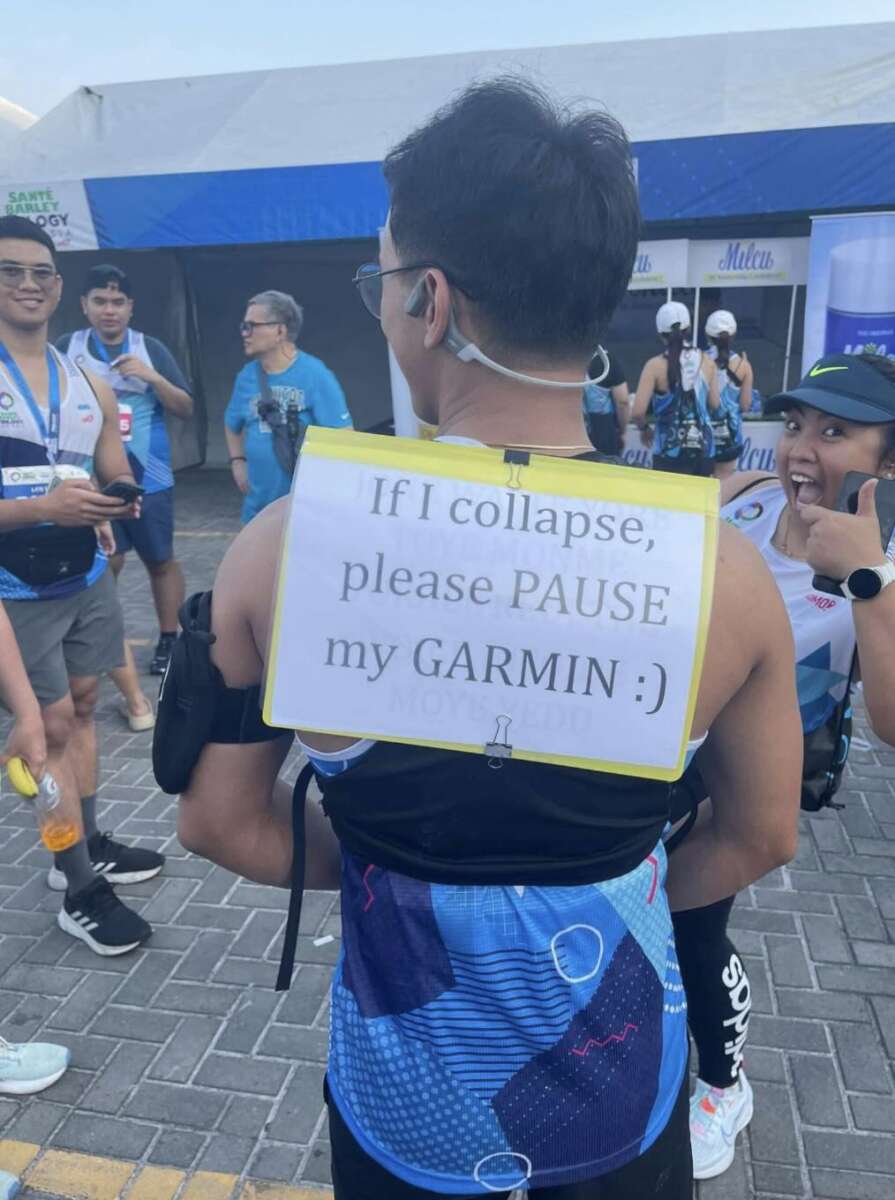 A runner at an outdoor event has a sign on their back that reads, "If I collapse, please PAUSE my GARMIN :)." Other people around are smiling and taking photos, with one woman giving a thumbs up.