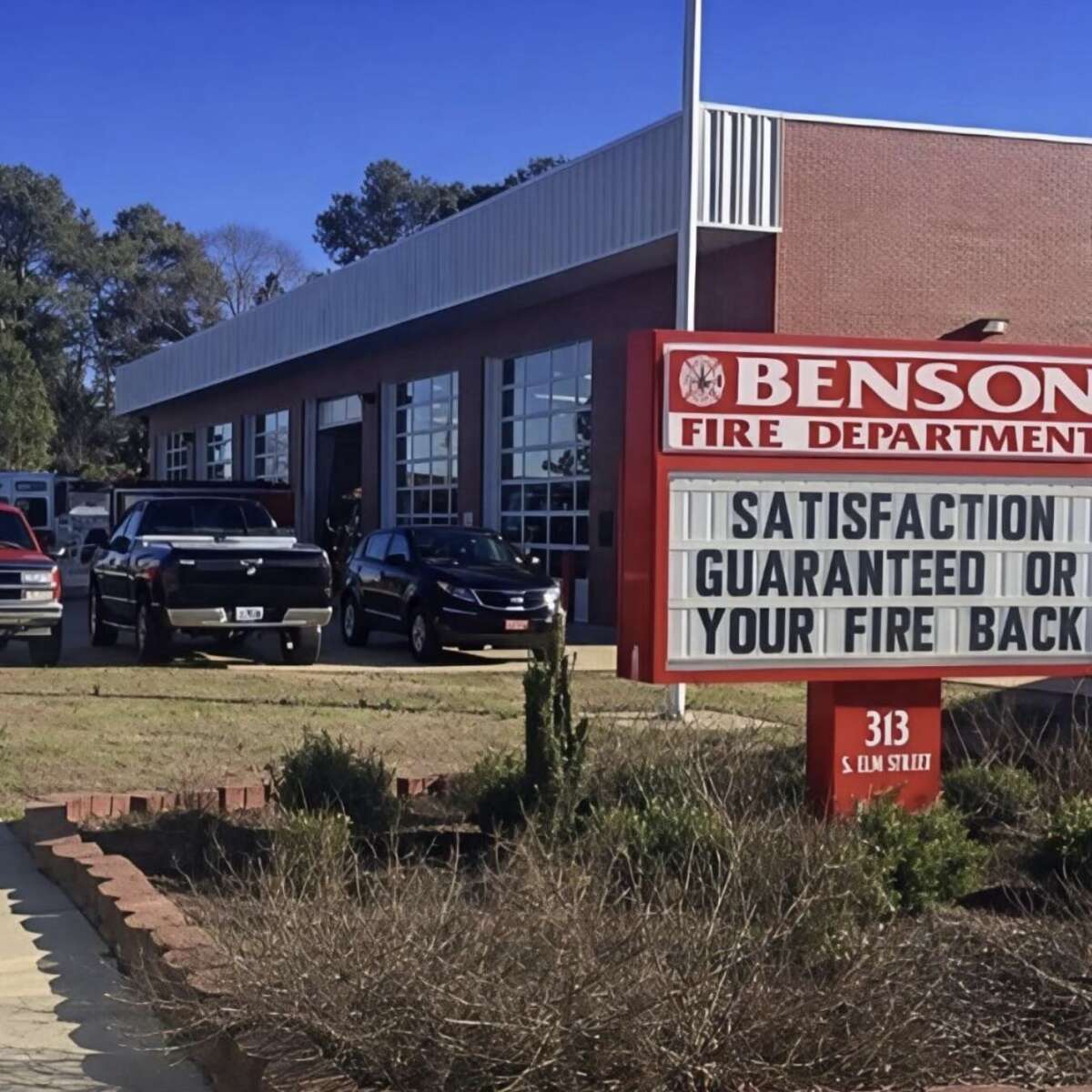 A fire station with several parked vehicles and a sign reading, "Satisfaction guaranteed or your fire back." The station is a red brick building with large windows. The address "313 S. Elm Street" is shown on the sign.