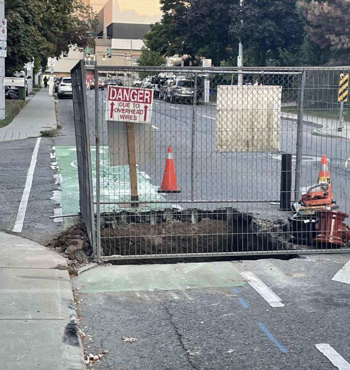 A fenced-off construction area blocks part of a sidewalk and bike lane. A "Danger: Due to overhead wires" sign is posted, with traffic cones and construction equipment nearby.