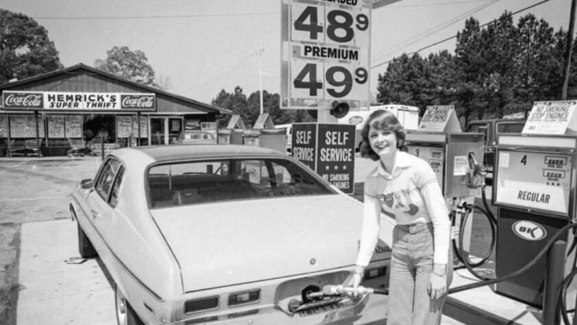 A woman smiles while pumping gas into a car at a vintage self-service gas station. A sign shows gas prices at 48.9 and 49.9 cents per gallon. A store named Hemrick’s Super Thrift is visible in the background.