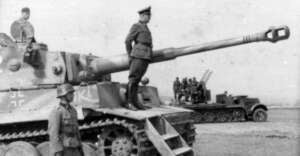 A black-and-white photo shows a uniformed officer standing on the turret of a German tank from World War II, with soldiers nearby and another military vehicle in the background.