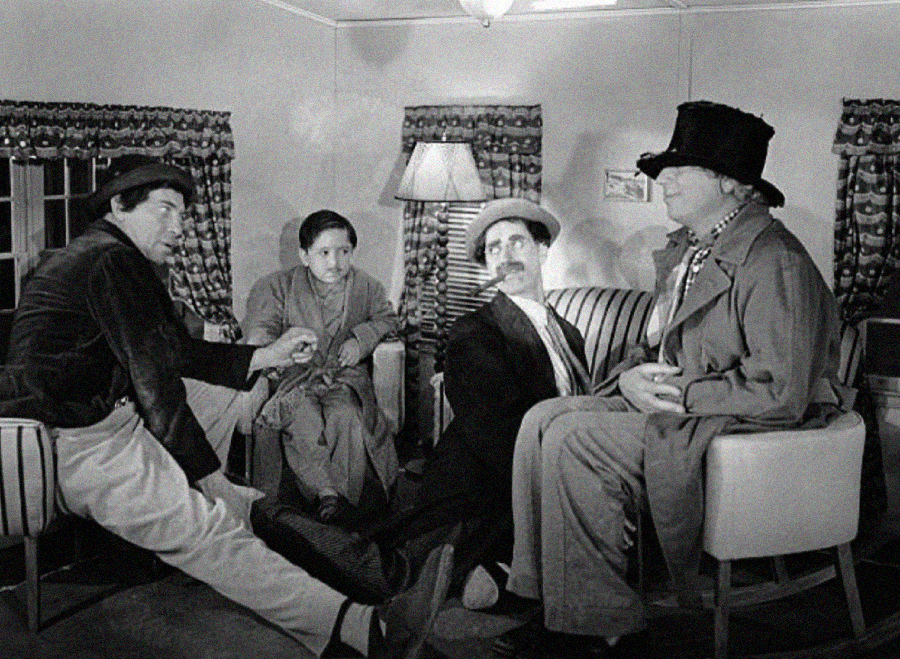 Four men in vintage clothing sit in a living room; two are seated in chairs, one wears a tall hat and coat, while the other two sit on the floor with their legs stretched out. The scene appears comedic and old-fashioned.