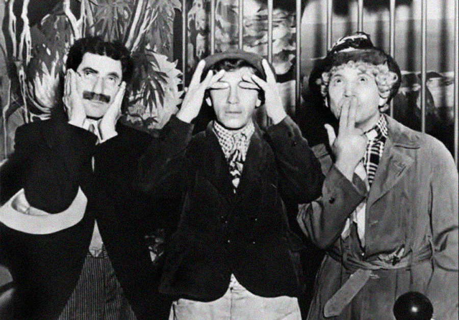 Three people dressed in old-fashioned costumes mimic “see no evil, hear no evil, speak no evil” poses, with one covering his eyes, another his ears, and the third his mouth, standing in front of a patterned background.