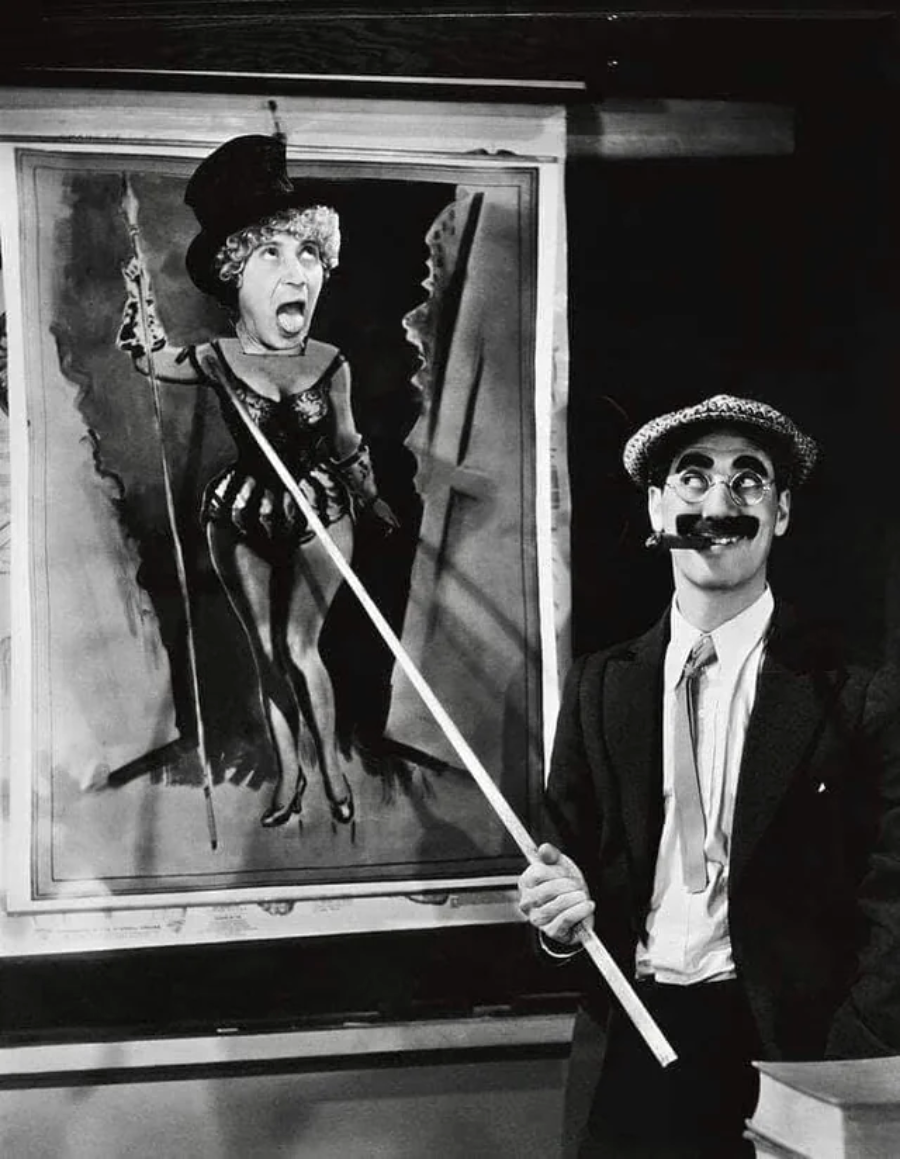 A man in a suit, fake mustache, and glasses points with a stick at a humorous caricature of a woman in a costume, drawn on a large sheet of paper.