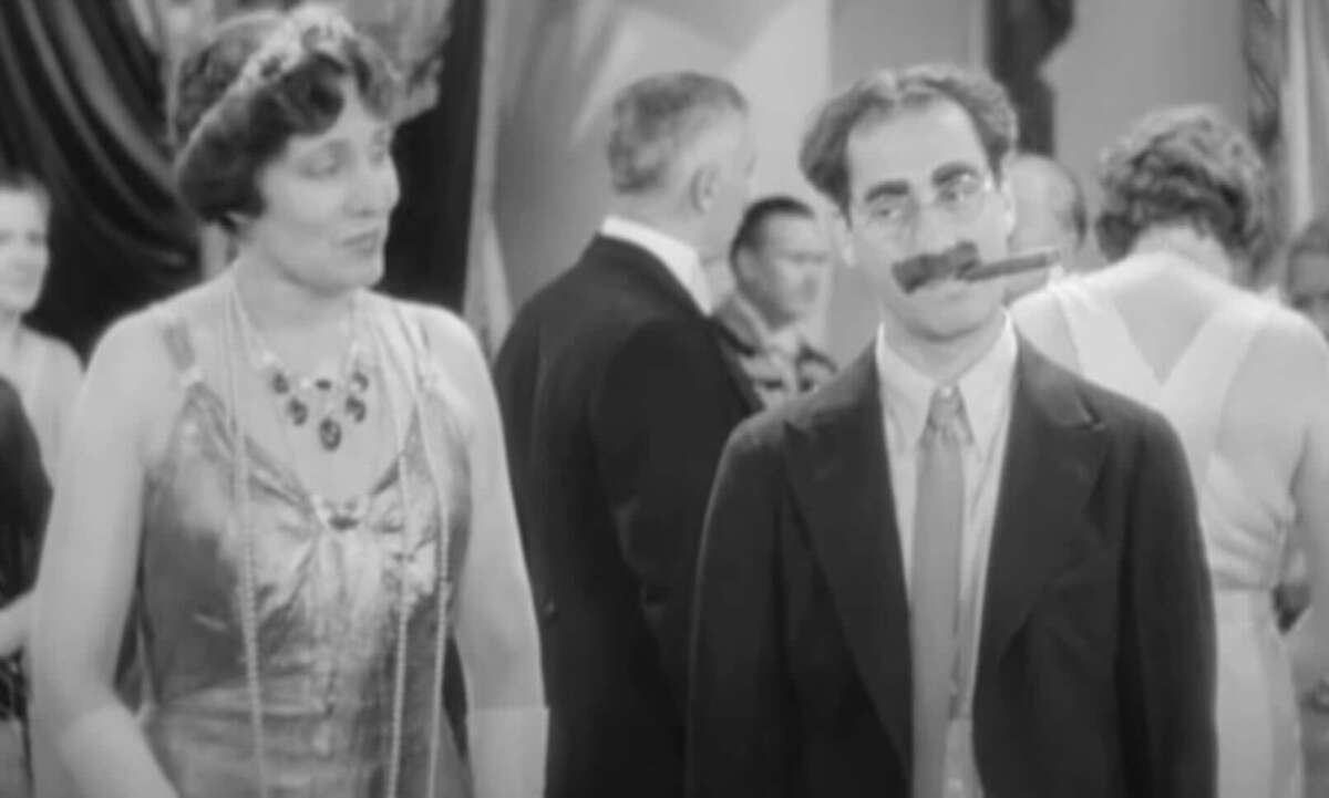 A woman in a beaded dress stands next to a man in a suit with a cigar in his mouth and glasses on, at a formal gathering with other people in the background.