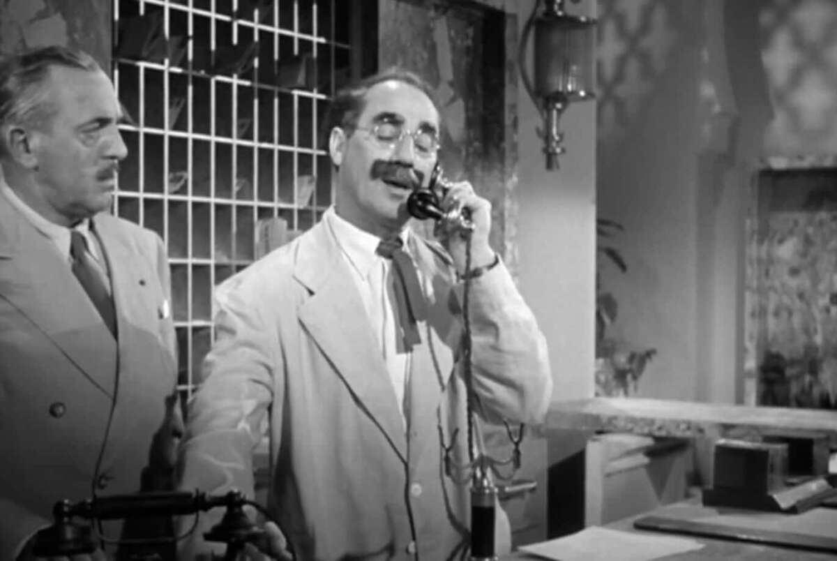 Two men in suits stand at a reception desk. The man in front, with glasses and a mustache, speaks on a vintage telephone. The background features a barred window and ornate decor. The scene is in black and white.