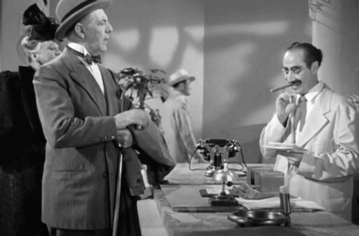 A man in a suit and hat stands at a counter, holding a jacket, while another man in glasses, a white coat, and a mustache holds a cigar and examines papers; vintage black-and-white scene.