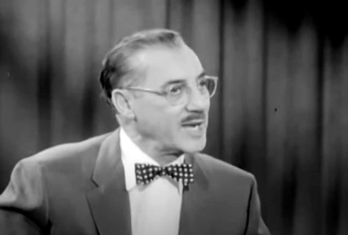 Black and white image of a man with glasses, neatly combed hair, and a mustache, wearing a suit and polka dot bow tie, speaking with an expressive look in front of a curtain background.