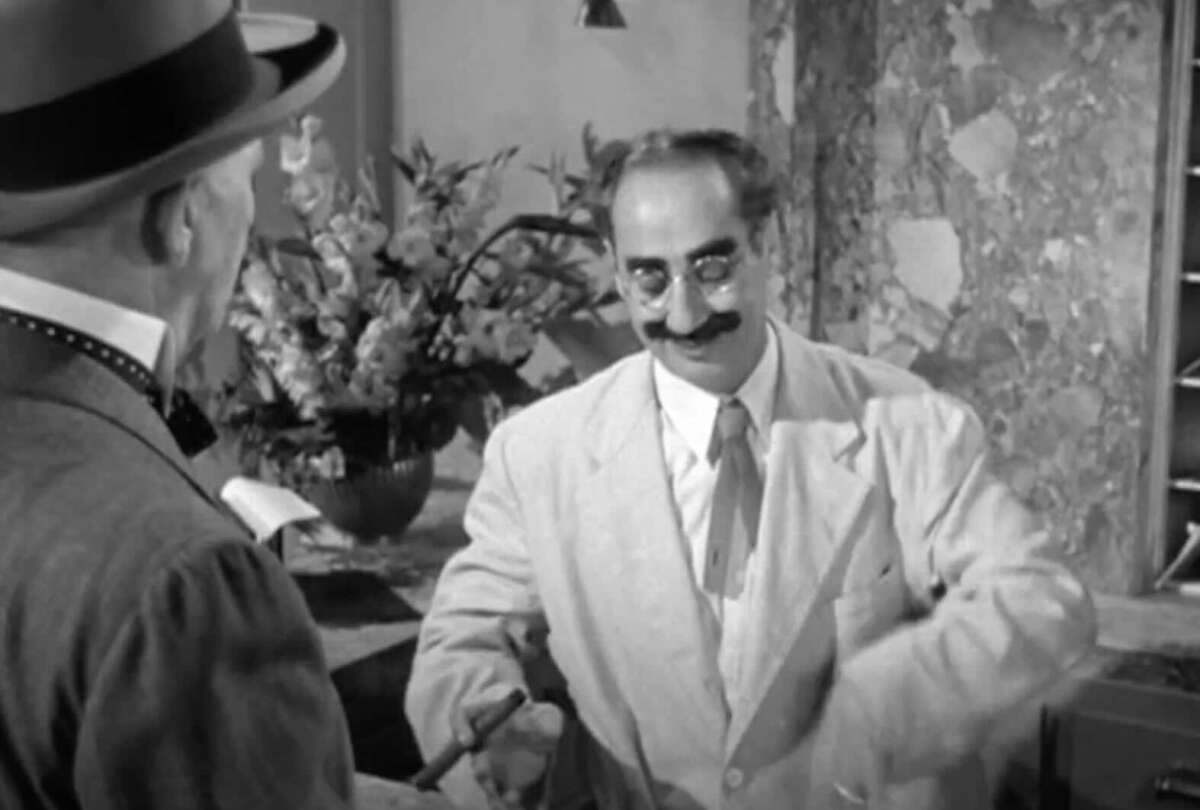 A man in a light suit and glasses with a thick mustache stands at a counter, smiling and gesturing with his hand, while another man in a hat and bowtie faces him. A vase of flowers is visible in the background.