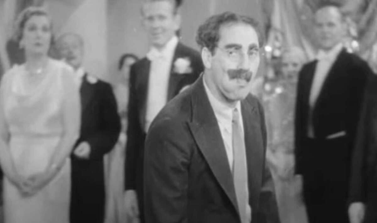 A man with thick eyebrows, glasses, and a mustache stands in the foreground at a formal gathering, surrounded by people in evening attire, in a black-and-white scene.
