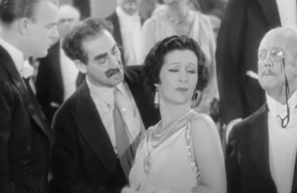A man with glasses and a mustache leans toward a stylishly dressed woman who looks away, while another man with a monocle stands nearby in a crowded, formal setting.