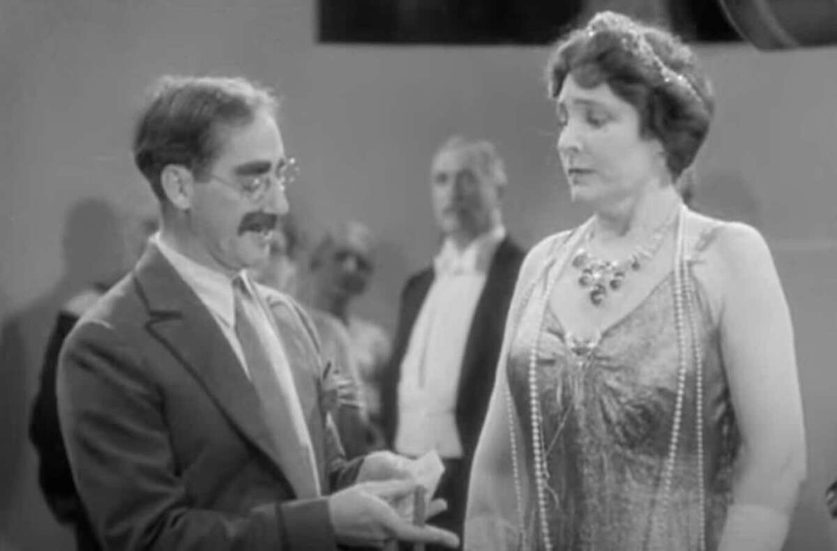 A man with glasses and a mustache gestures with his hands while speaking to a woman in a beaded dress and layered necklace at a formal event, with other people blurred in the background.