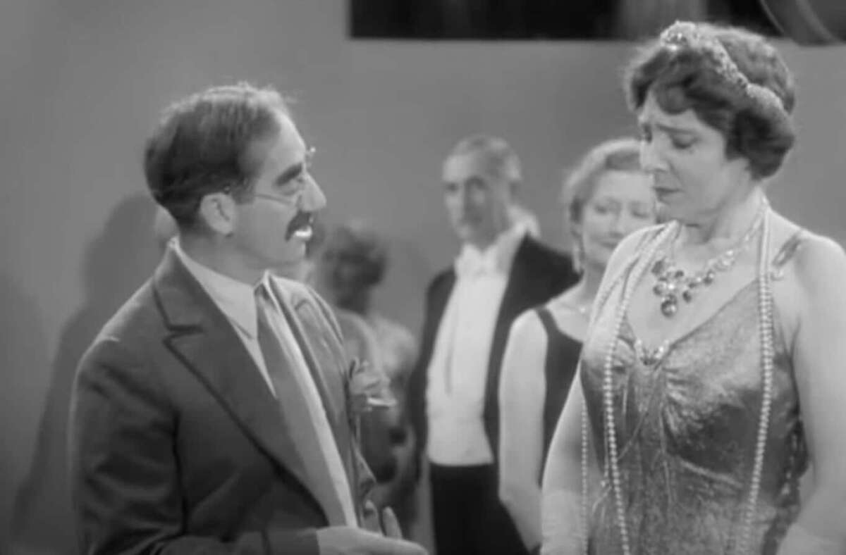 A man with a mustache in a suit speaks to a woman wearing a beaded dress, pearl necklace, and headpiece at a formal gathering, with other people blurred in the background.