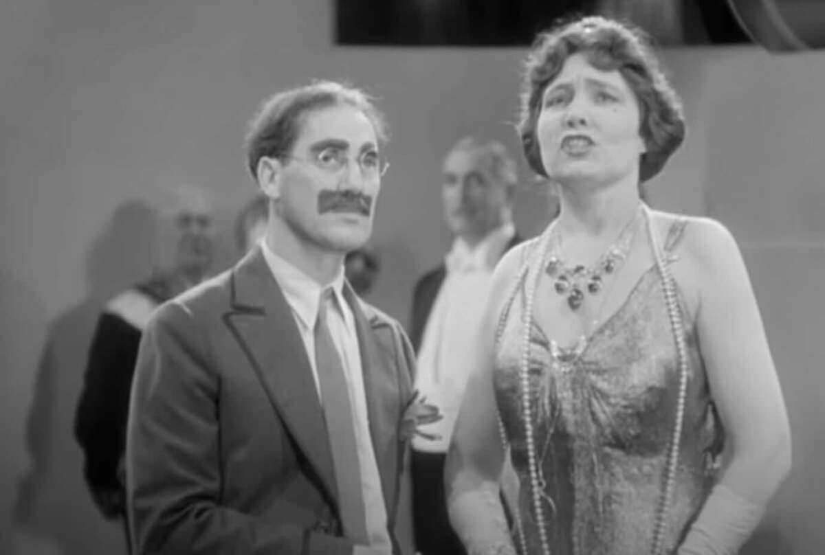 A man with glasses, a painted mustache, and a suit stands next to a woman in a sparkling dress and necklace. Both are indoors at an event, with people visible in the blurred background.