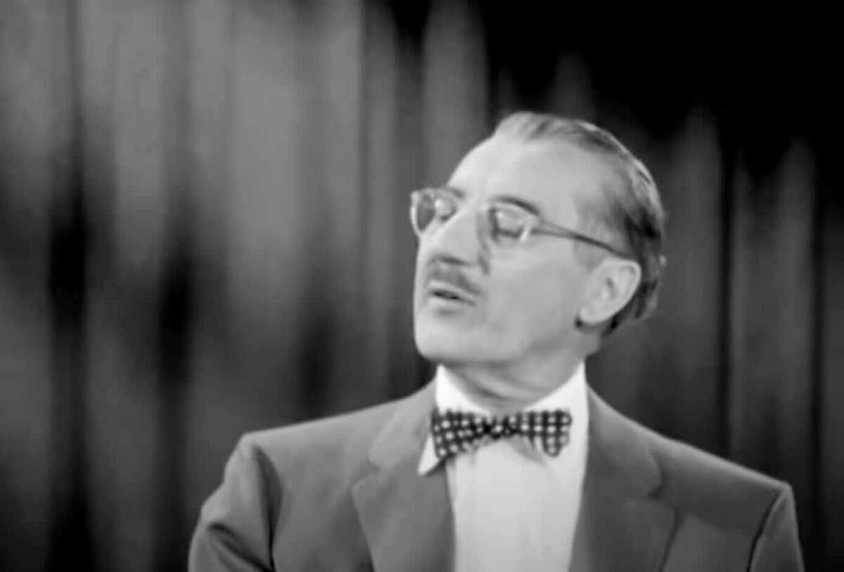 A man with glasses and a mustache wearing a bow tie and suit looks to his left. The background features vertical, dark curtains in an indoor setting. The image is in black and white.