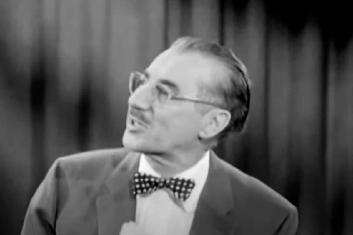 A man with glasses, slicked-back hair, and a mustache wears a polka-dot bow tie and suit, looking to the side in front of dark curtains. The image is in black and white.