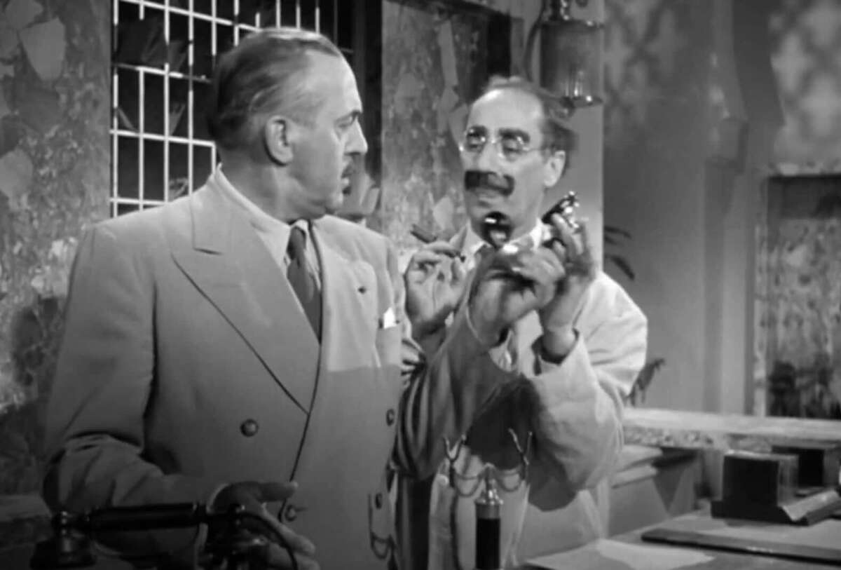 Two men in suits stand indoors; one man, wearing glasses and a fake mustache, gestures animatedly with his hands while the other looks at him with surprise. A barred window and marble walls are in the background.