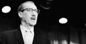 A man with glasses, a mustache, and slicked-back hair wears a suit and tie, standing under bright stage lights in front of a curtain. The image is in black and white.