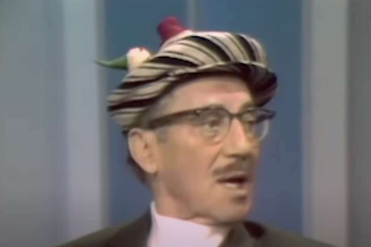 An older man with glasses and a mustache wears a striped hat with a feather. The background is blue and gray vertical panels.