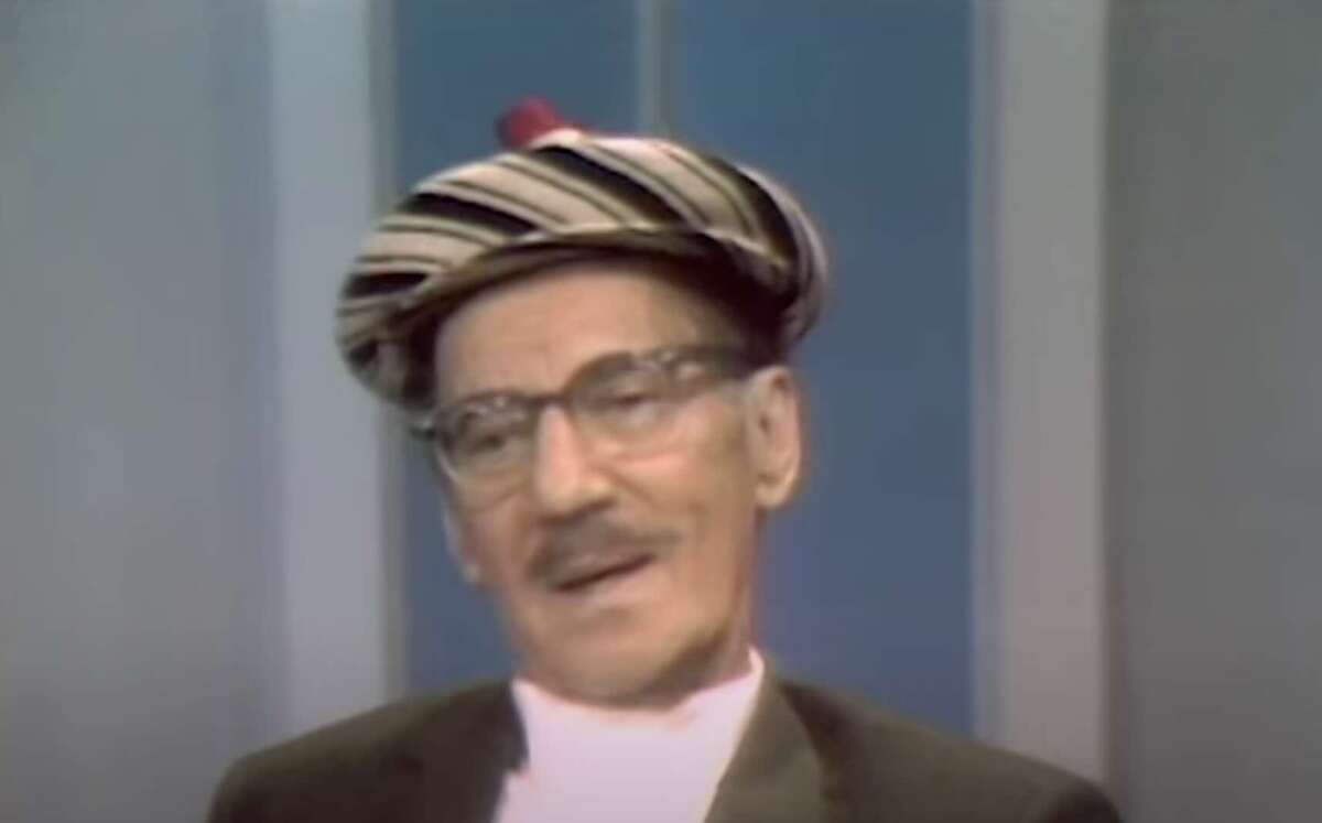 An older man with a mustache, glasses, and a striped beret is sitting and smiling softly in front of a blue and gray background. He is wearing a suit and appears to be on a television set.