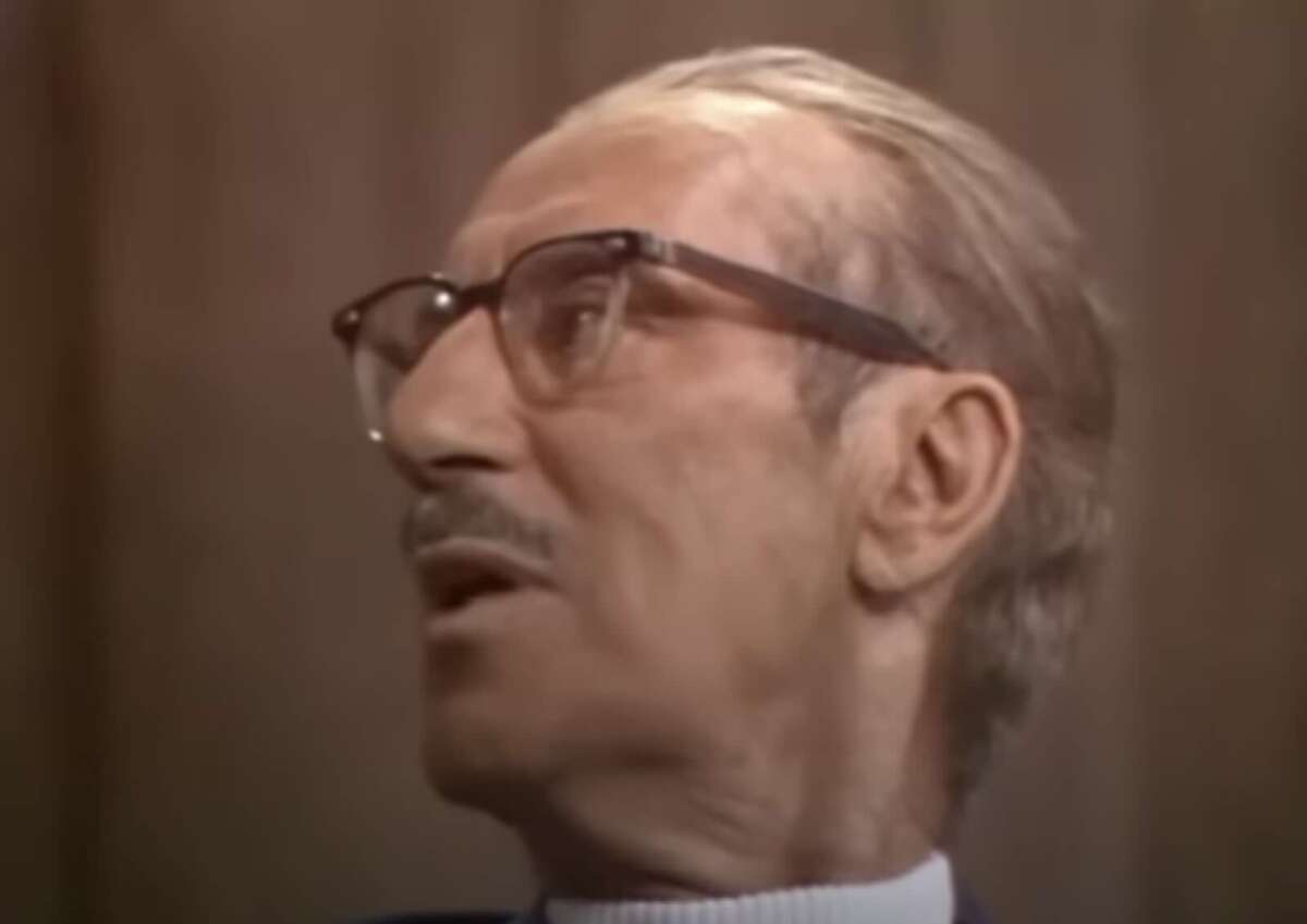 An elderly man with glasses and slicked-back hair, wearing a mustache and a suit, is looking to his left against a plain, brown background.