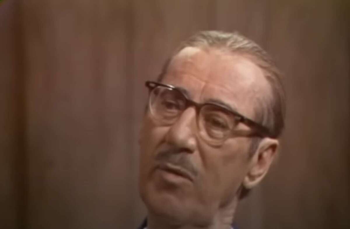 An older man with slicked-back hair, glasses, and a mustache looks slightly to the side with a thoughtful expression, set against a plain brown background.