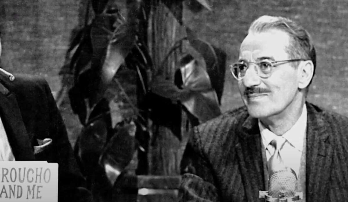 A man with glasses, mustache, and striped suit sits at a desk with a microphone. A book titled "Groucho and Me" is partly visible. Large leafy plants are in the background. The image is in black and white.