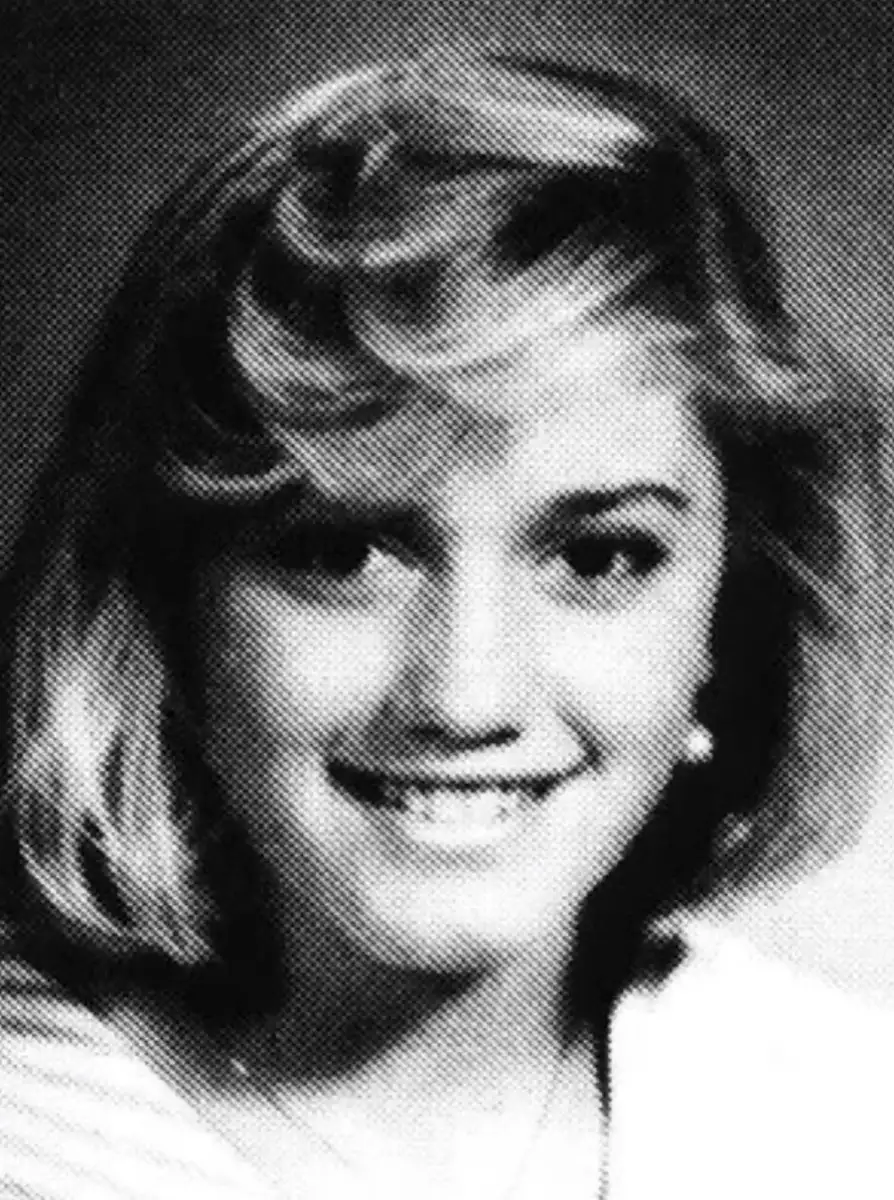 Black and white portrait of a young woman with short, wavy blonde hair, smiling at the camera. She is wearing earrings and a light-colored top. The image has a grainy, yearbook-style quality.