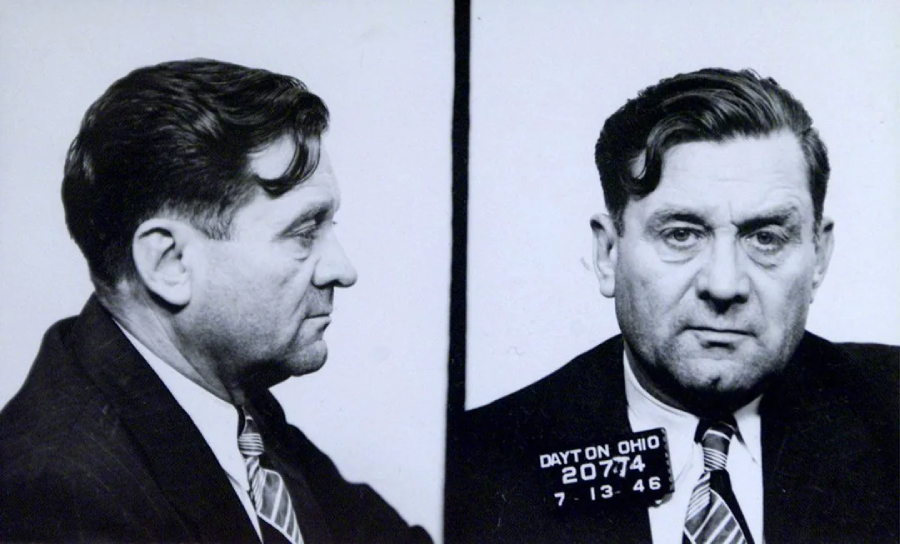 Black and white mugshot of a middle-aged man with wavy dark hair, wearing a suit and tie. There are two views: a side profile and a front-facing shot. A placard with identification numbers and a date is visible.
