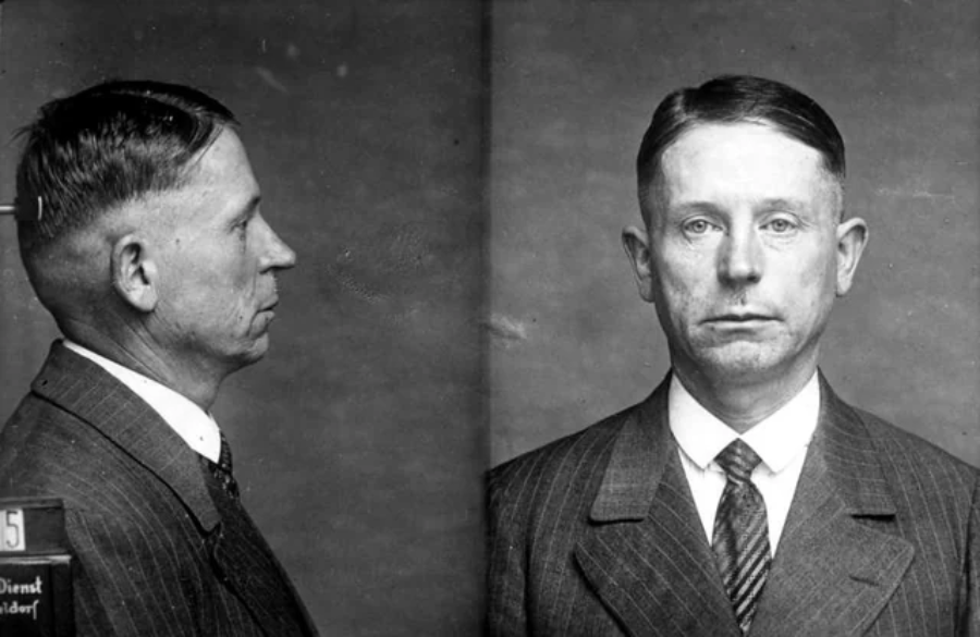 Black and white mugshot of a man in a suit and tie, shown from the front and side against a plain background. The man has short, neatly parted hair and a serious expression.