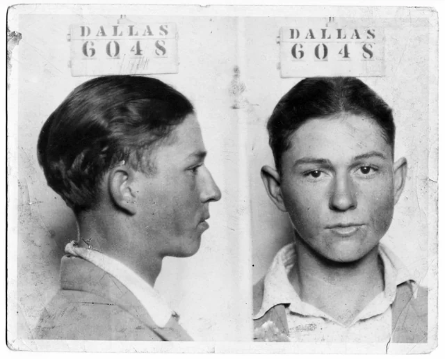 Black and white mugshot photo showing a young man in two views: profile and front-facing. He wears a collared shirt and jacket. Above each image is a sign reading "DALLAS 6048." The photo appears old and worn.